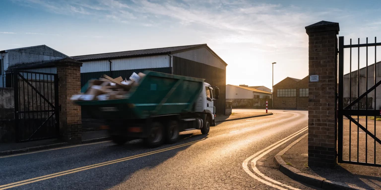 A skip wagon carrying a full chain-lift skip pulling out of an industrial estate gate at dawn, wheels blurred with motion