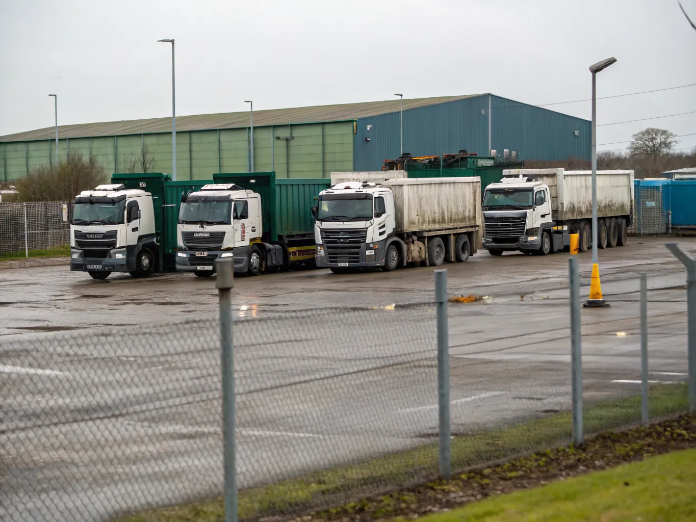 Row of waste collection lorries and skip trucks parked in a fenced haulage depot yard on wet tarmac