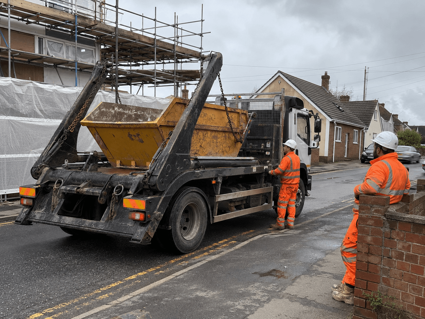 A driver in yellow hi-vis standing beside a hook-loader wagon in a UK depot yard, with a chain-lift skip and stacked containers behind